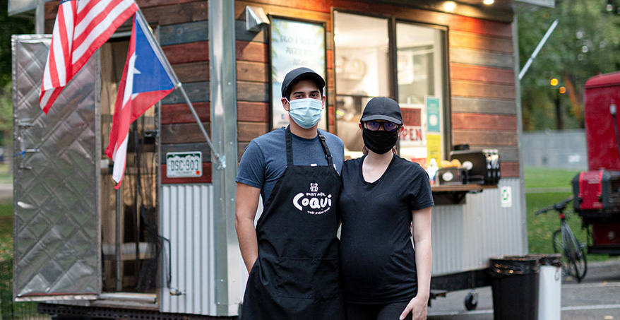 two people wearing face masks standing in front of a food truck
